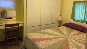 Un dormitorio con cama, escritorio con silla, armario, ventilador y ventana con cortinas.