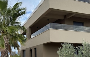 Un immeuble moderne à plusieurs étages, doté de balcons, d’un palmier et d’un petit arbuste.