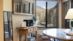 A room with a large window offering a mountain view, a wooden console table with speakers and a lamp, and a round glass table. - Fallridge Condo - Close to Bus & Nordic Center (Vail)