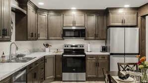 A modern kitchen with wooden cabinets, stainless steel appliances, a marble countertop, and a dining area with a table set for two. - Farm Views | 10min to Sight & Sound (Ronks)