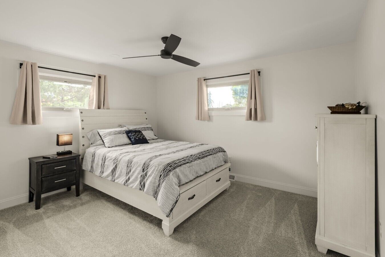 A bedroom with a bed, nightstand, lamp, ceiling fan, windows, and a wardrobe.