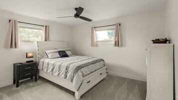 A bedroom with a bed, nightstand, lamp, ceiling fan, windows, and a wardrobe.