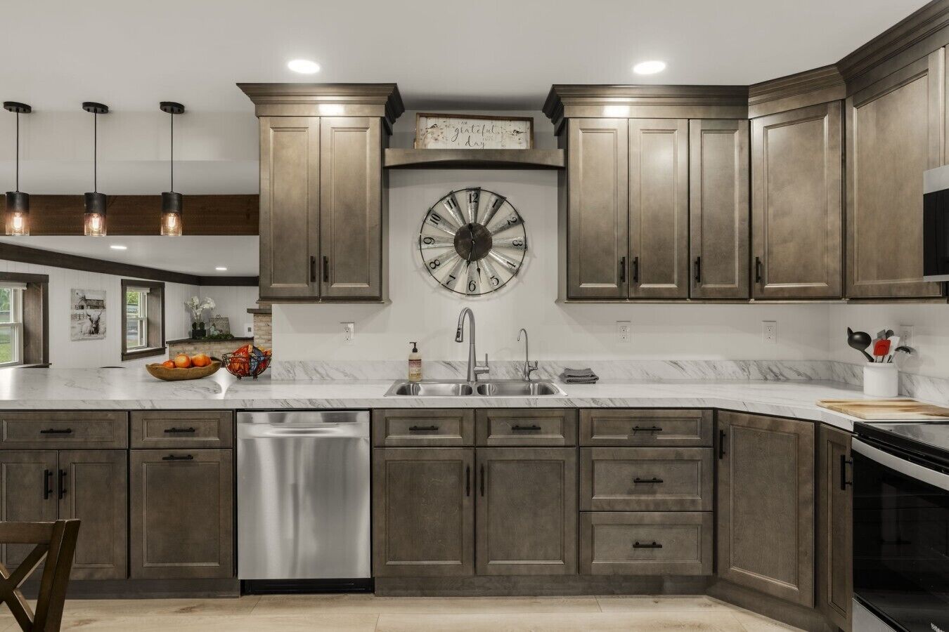 A kitchen with dark wood cabinets, a stainless steel dishwasher, a large wall clock, and pendant lighting.