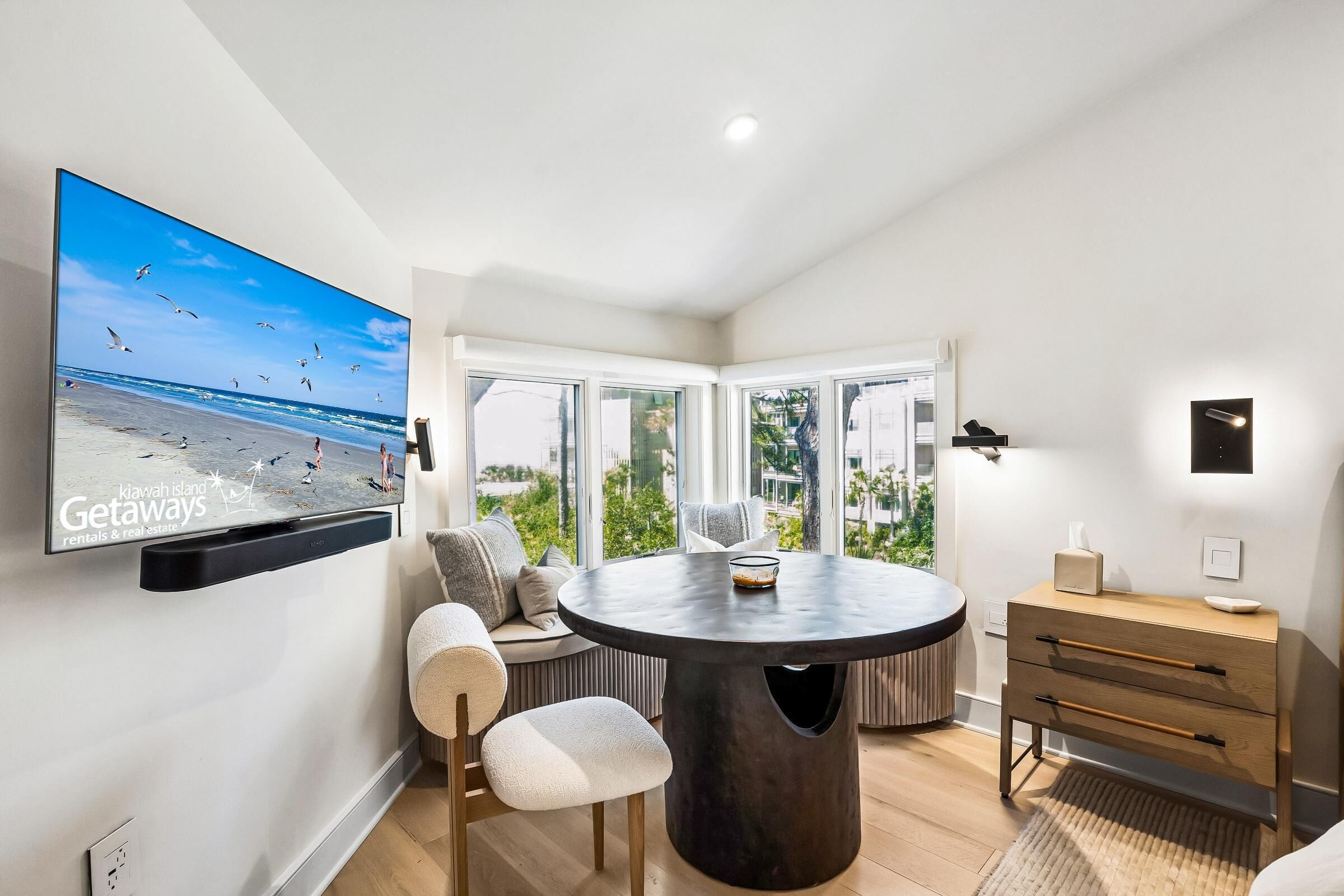Een moderne kamer met een grote tv waarop een strandscène wordt getoond, een ronde eettafel, een bank en een houten dressoir.