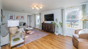 A living room with a sofa, coffee table, TV, and a cabinet. There is a window with curtains and a view of trees.
