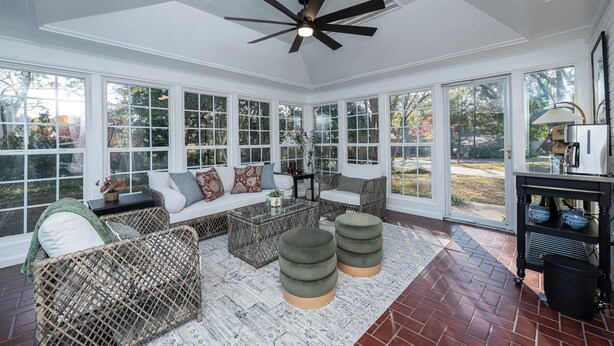 A sunroom with a ceiling fan, large windows, wicker furniture, and a brick floor.