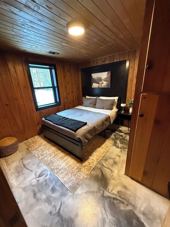 A wooden-paneled bedroom with a bed, a nightstand, a window, and a picture on the wall.