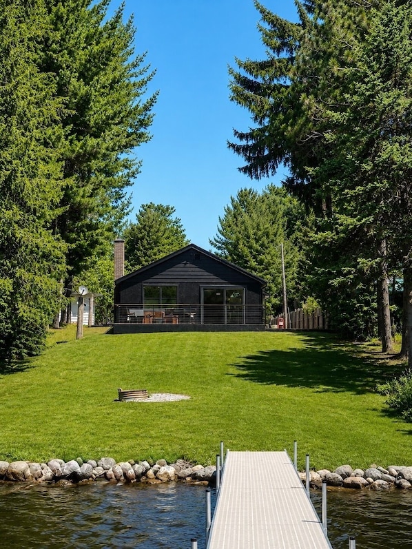 A modern cabin with a dock extending into a lake, surrounded by dense trees. - Waterfront- Modern Luxurious Cabin on Clam Lake – Torch Lake Boating (Bellaire)