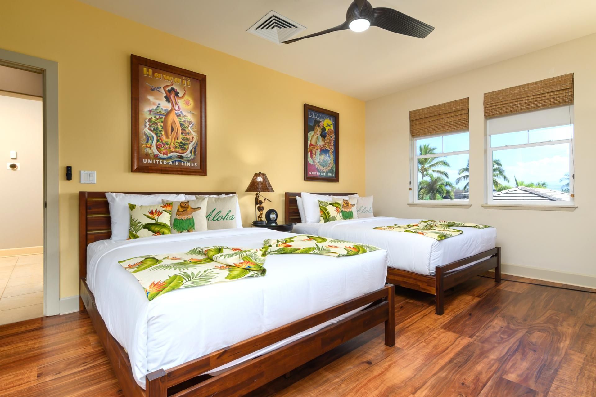 A hotel room with two wooden beds, a ceiling fan, and two windows with views of palm trees.