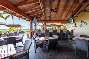 Un espace extérieur aménagé en terrasse, avec des tables et des chaises en bois, un ventilateur de plafond et une vue sur l’océan.
