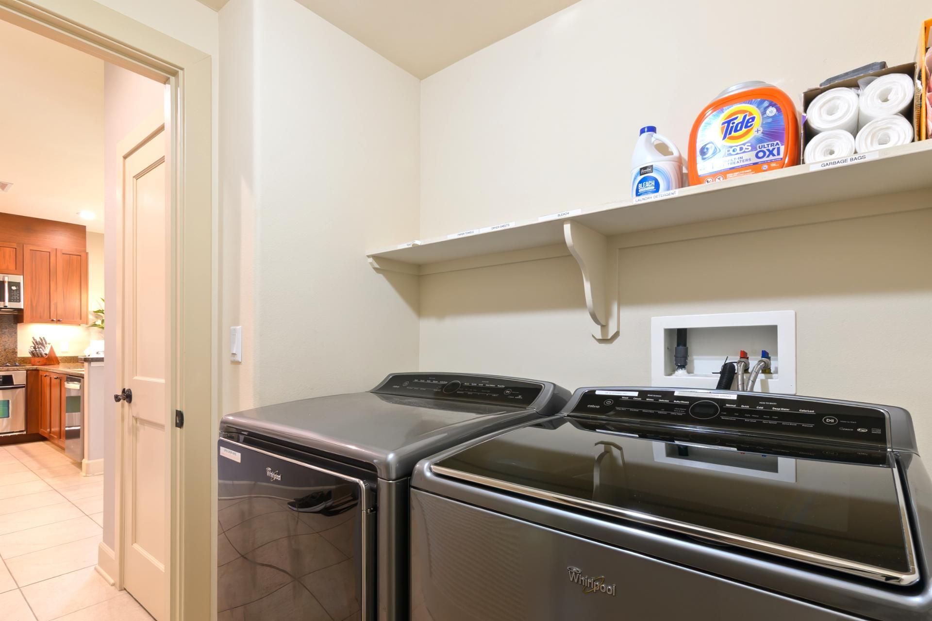 A laundry room with a Whirlpool washer and dryer, a shelf with Tide detergent, and a microwave in the adjacent kitchen.