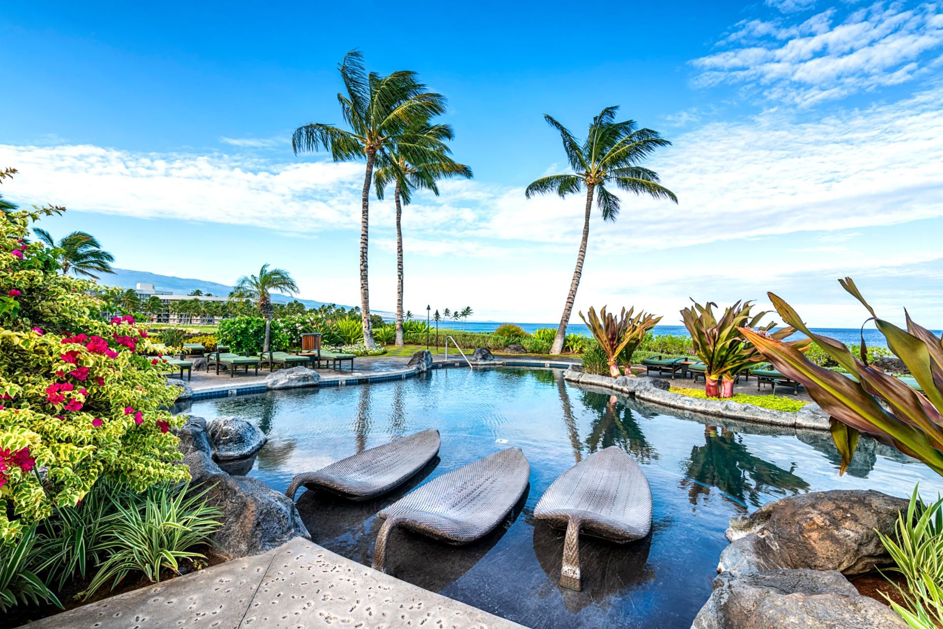 A pool with lounge chairs, palm trees, and a view of the ocean.