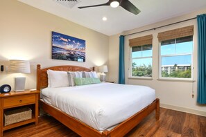 A bedroom with a wooden bed, a nightstand, a wall-mounted lamp, a ceiling fan, and a large window with curtains. - Halii Kai | Ocean View 2 BR | Car Incl. w/ 6+ Nights | HLI-7A by KBM (Waikoloa)