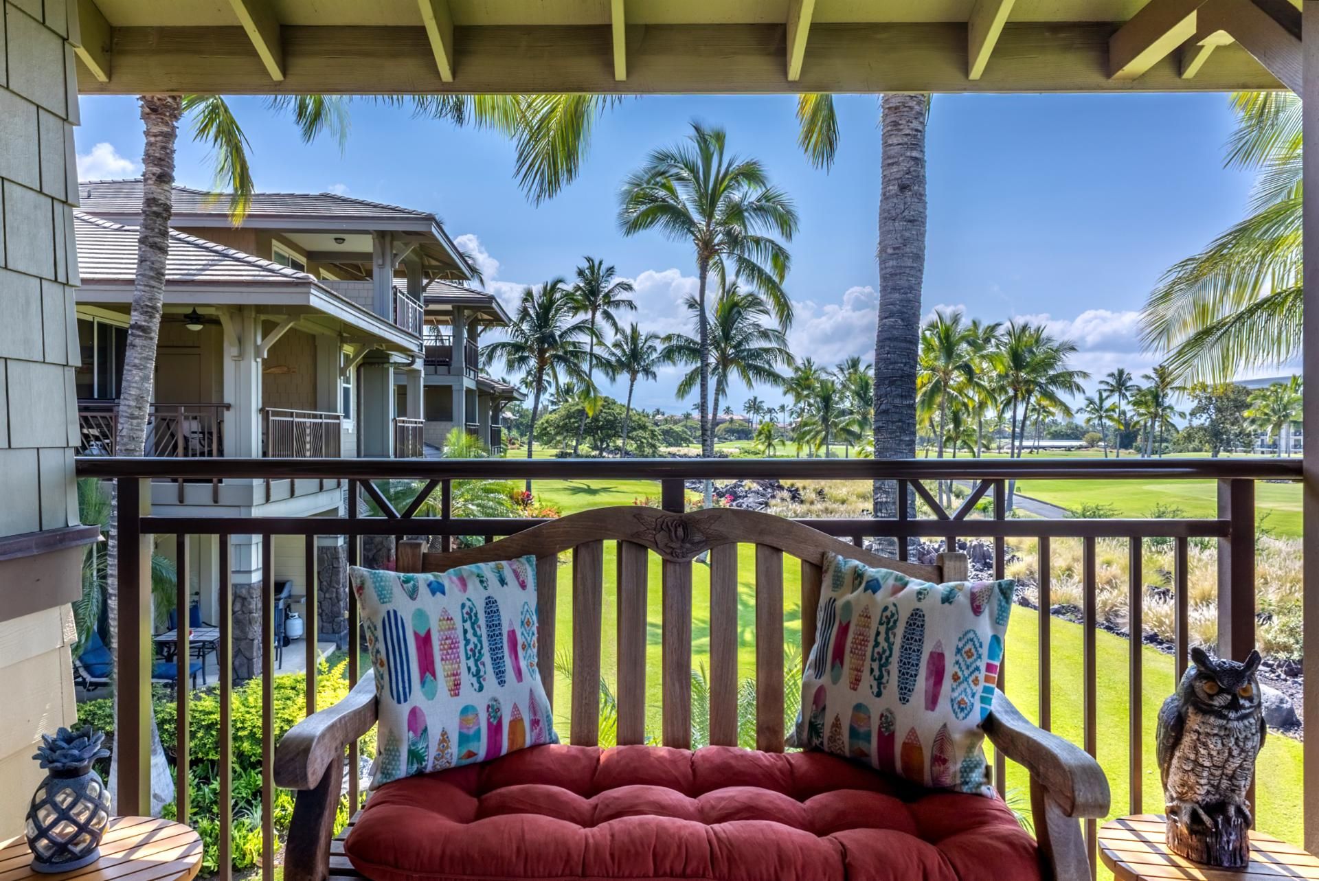 A balcony with a wooden bench, a decorative owl statue, and a view of a house and palm trees.