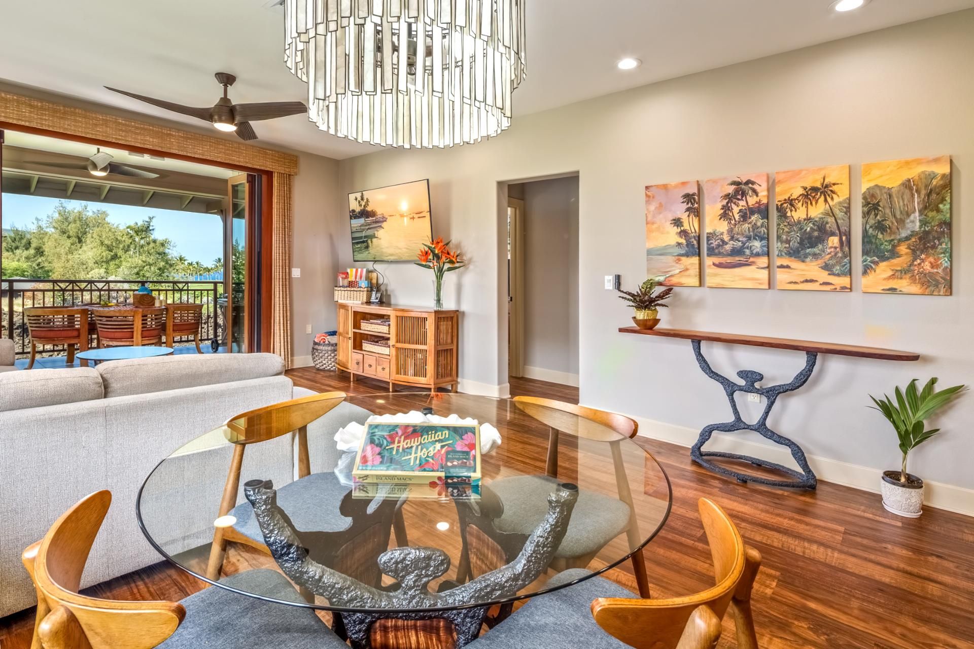 A modern living room with a glass coffee table, wooden chairs, a sofa, a ceiling fan, and artwork on the walls.