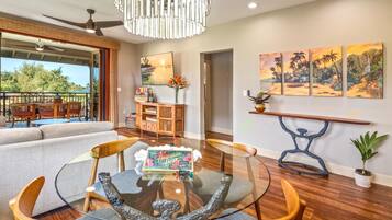 A modern living room with a glass coffee table, wooden chairs, a sofa, a ceiling fan, and artwork on the walls.