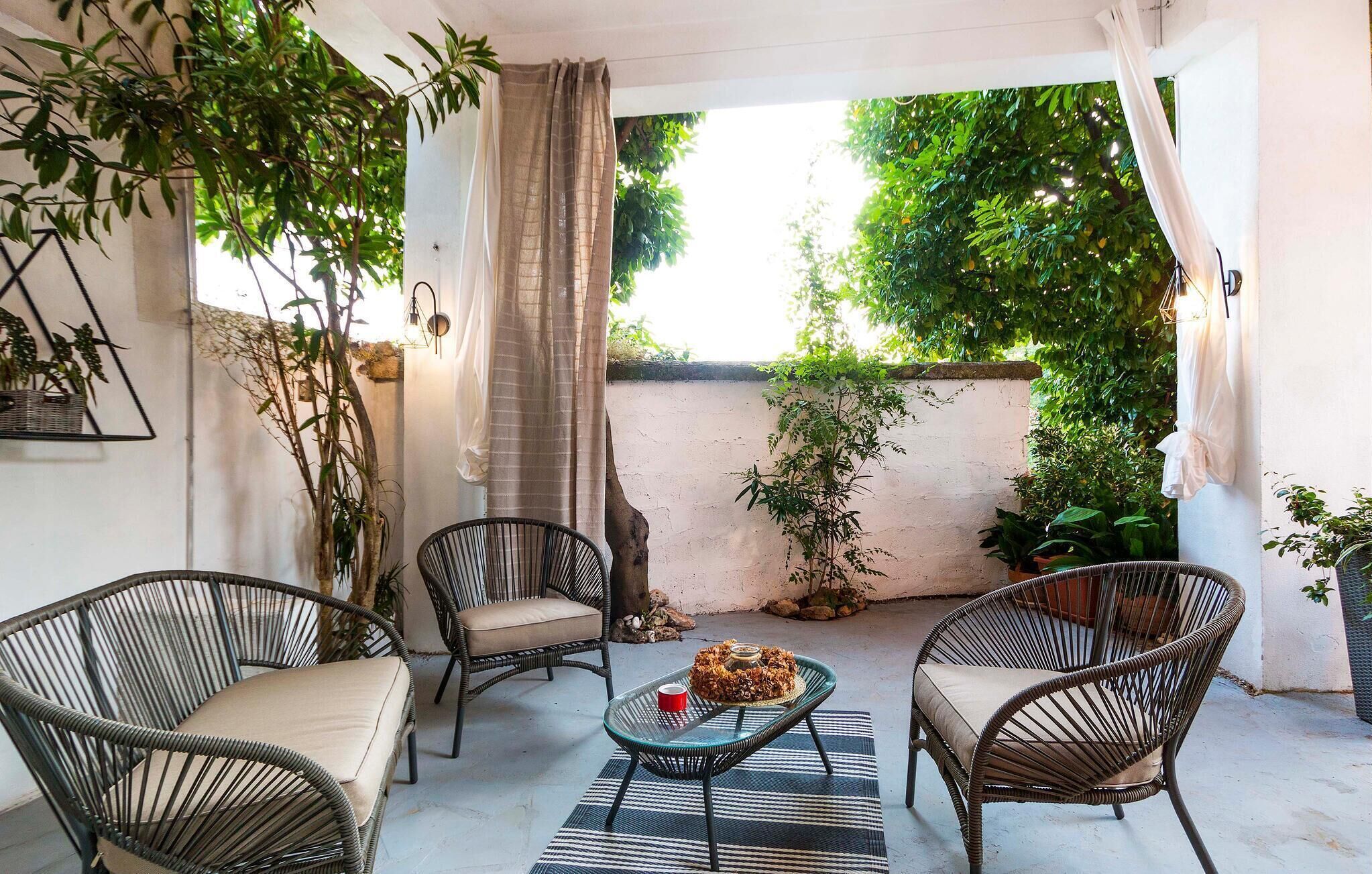 Une terrasse avec des fauteuils en osier, une table en verre et une vue sur la verdure, visible à travers un mur blanc.