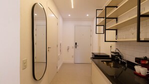 A modern kitchen with a black countertop, a large mirror, and a white door in the background. - Studio com Varanda a 280m do Metr Vila Madalena (São Paulo)