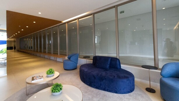 A modern lobby with a curved blue sofa, two round tables with potted plants, and a glass wall with a view of the interior. - Studio com Varanda a 280m do Metr Vila Madalena (São Paulo)