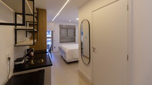 A modern hotel room with a kitchenette, a bed, a large mirror, and a door leading to another room. - Studio com Varanda a 280m do Metr Vila Madalena (São Paulo)