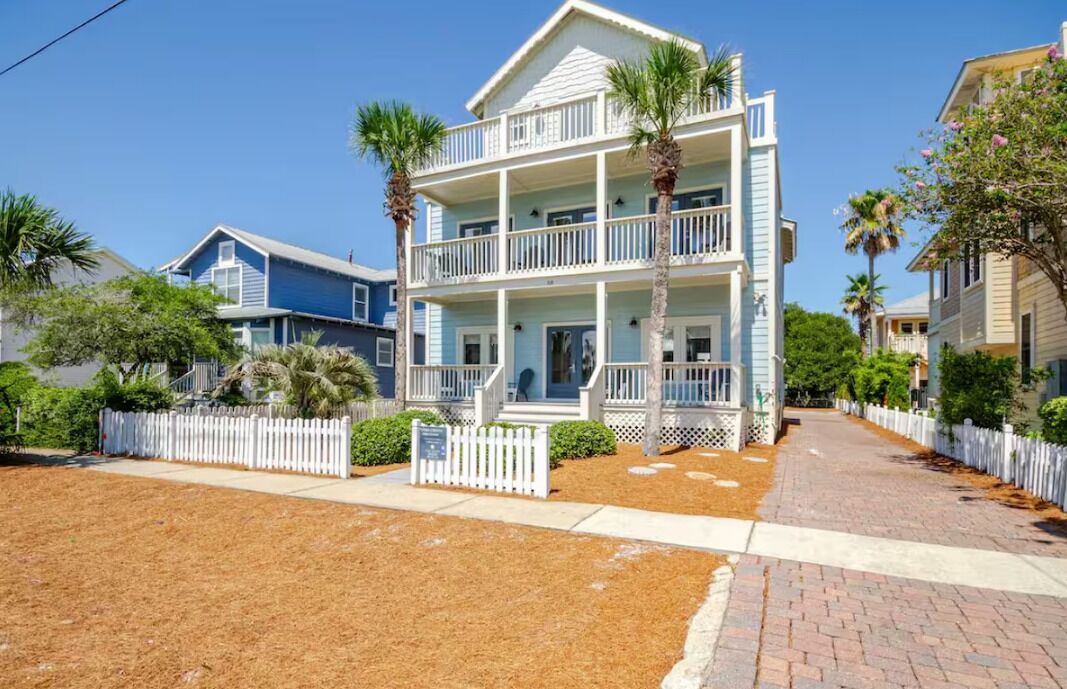 A two-story beachfront house with a balcony, surrounded by a white picket fence and palm trees.