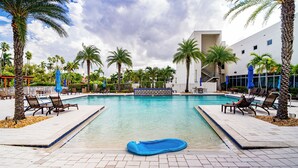 Ein Swimmingpool mit Liegestühlen und Sonnenschirmen, umgeben von Palmen und einem modernen Gebäude.