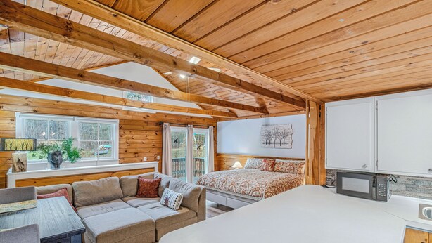 A cozy cabin interior with a wooden ceiling, a sofa, a bed, a dining table, and a kitchen area.