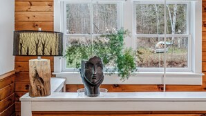 A room with wooden paneling, a white window with a view of trees, a table with a sculpture, and a lamp with a tree pattern.