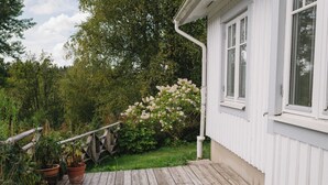 Puinen terassi johtaa valkoiseen rakennukseen, jossa on suuret ikkunat. Rakennuksen ympärillä on vehreää kasvillisuutta ja kukkivia kasveja.