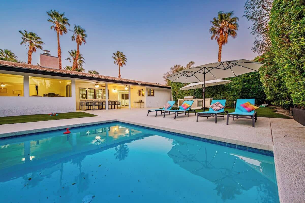 A swimming pool with lounge chairs and umbrellas, surrounded by palm trees and a house with a patio.