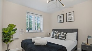 Un dormitorio con cama, ventana, radiador, planta y cuadros en la pared.