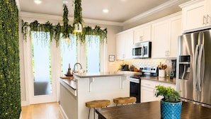 Une cuisine moderne avec des meubles blancs, des appareils en acier inoxydable, un îlot central doté de tabourets et une plante suspendue servant de décoration.