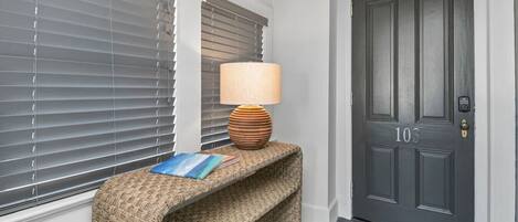 A dark grey door with the number 103, a woven bench with a lamp and a book, and grey blinds.