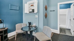 A room with a blue wall, a white shelf with books, a coffee machine, two chairs, and a shower area. - Walk to Coffee, Dining & Campus | Historic Studio (Columbia)