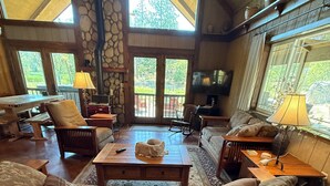 Uma sala de estar ampla, com um sofá grande, uma mesa de centro e uma lareira. Várias janelas oferecem luz natural e vista para o exterior.