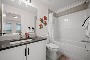 Une salle de bain moderne avec un lavabo, des toilettes et une baignoire, tous de couleur blanche. On y trouve également des éléments décoratifs muraux et un rideau de douche à motifs.