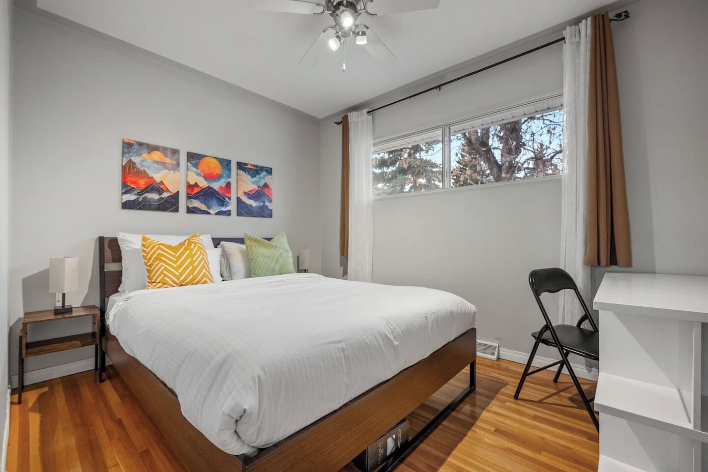 A modern bedroom with a large bed, a desk, and a ceiling fan. There are three paintings on the wall and a window with curtains.