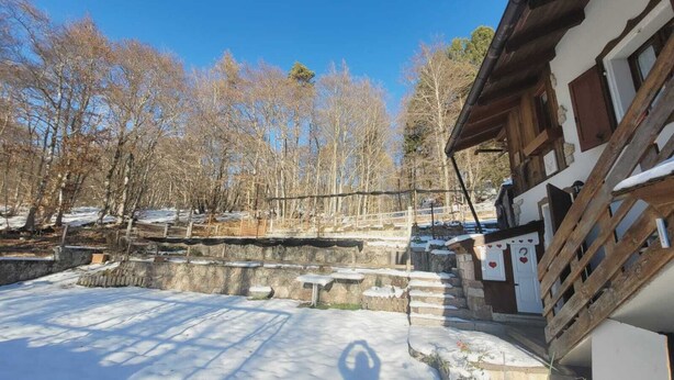 Un chalet rustique aux murs de pierre, doté d’une terrasse en bois et entouré d’un paysage enneigé.