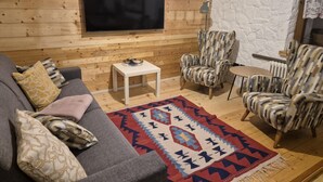 Un salon chaleureux avec un mur en bois, un téléviseur à écran plat, un canapé gris agrémenté de coussins à motifs, deux fauteuils à motifs, une petite table blanche et un tapis coloré.