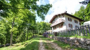 Une maison de campagne au cachet rustique, avec un mur en pierre et une clôture en bois, entourée d’une végétation luxuriante et bordée d’un chemin de gravier.