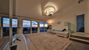 A modern living room with a large sliding glass door leading to a balcony, a white sofa, a glass coffee table, and a large window with a view of the sunset. - Gorgeous home in Podstrana (Podstrana)