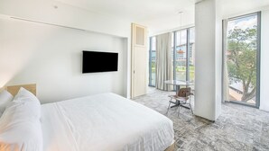 A modern hotel room with a large bed, a flat-screen TV mounted on the wall, a small table with a chair, and a large window offering a view of the city.