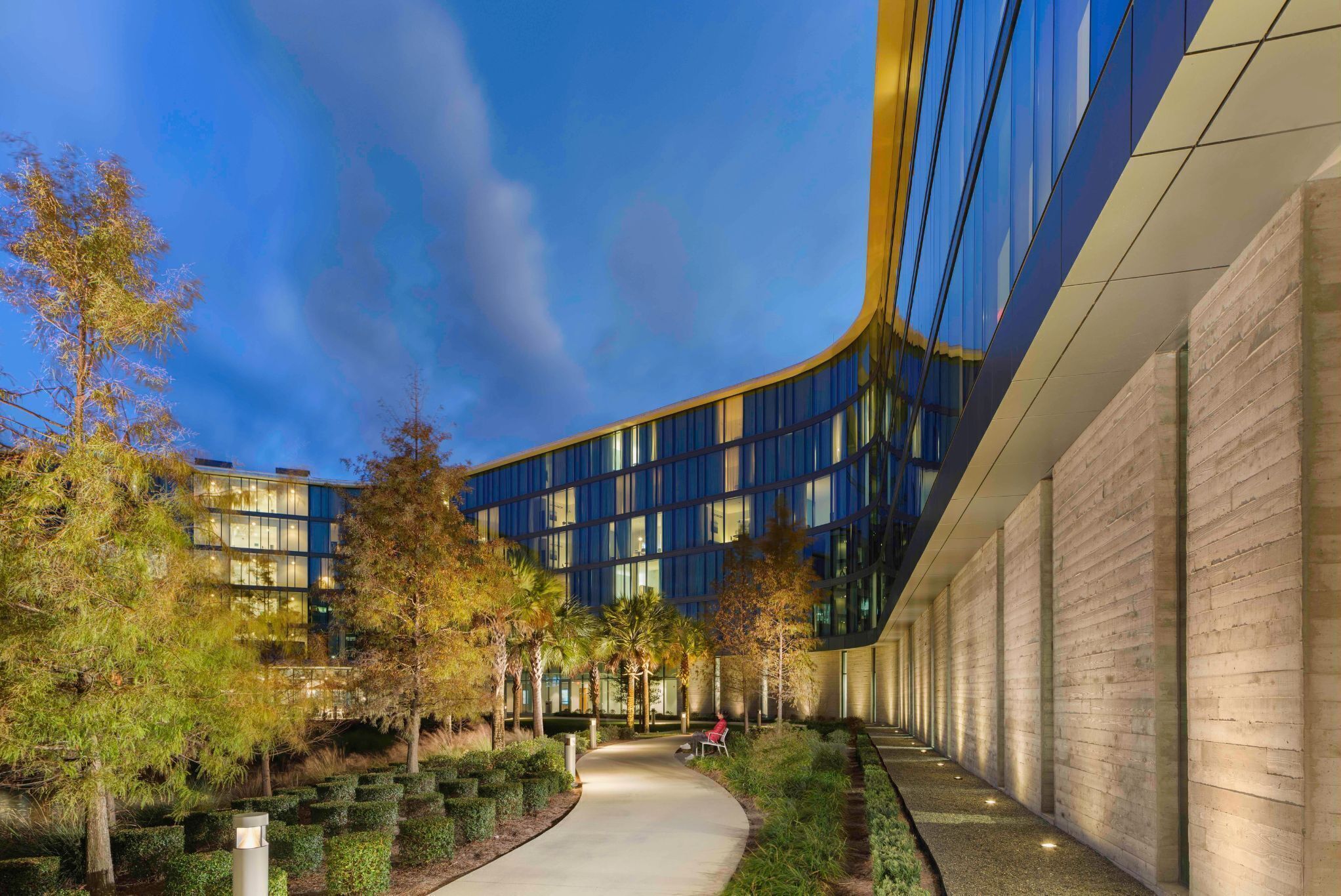 A modern building with a curved glass facade, surrounded by a landscaped garden with pathways and trees.