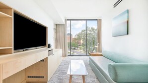A modern living room with a flat-screen TV, wooden shelving, a small white table, a green sofa, and a large window offering a city view.