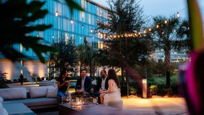 Een terras buiten met een vuurkorf, lichtjes en een modern gebouw op de achtergrond.