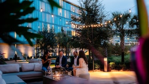 Un espacio al aire libre con una fogata, luces colgantes y un edificio moderno al fondo.