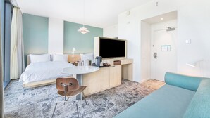 A modern hotel room with a bed, a desk with a chair, a TV, and a sofa.