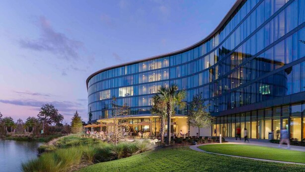 A modern hotel with a curved glass facade, outdoor seating area, and a small pond.