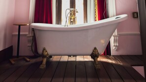 A freestanding bathtub in a room with wooden flooring and a window with red curtains. - La Casa Rosa Imperial (Mérida)