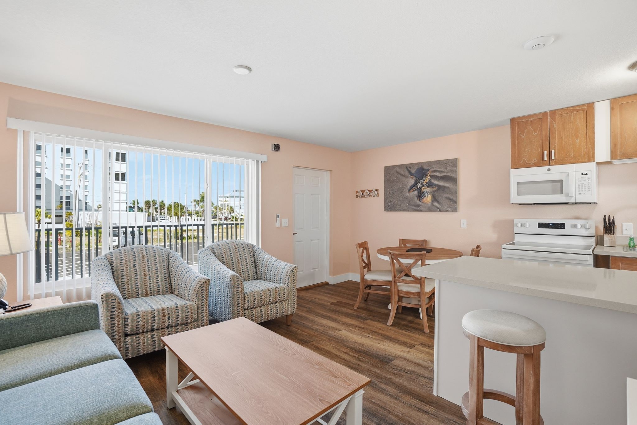 A living room with a sofa, a dining table with chairs, a kitchen area with a microwave and stove, and a balcony with a view of buildings.